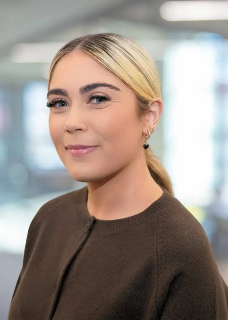 Blonde woman in a ponytail and brown top smiles in an office.
