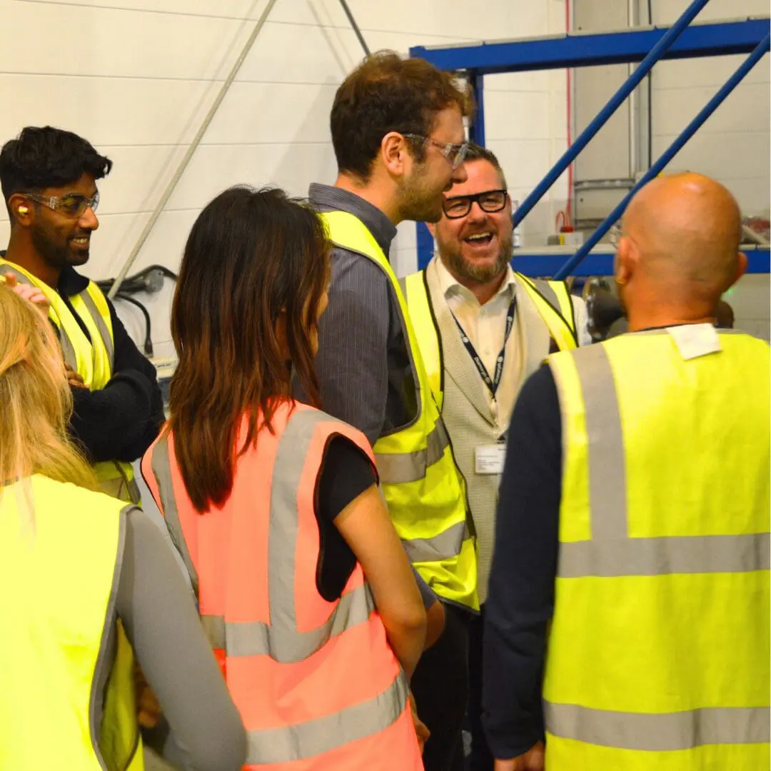 People in safety vests talk and laugh together in an industrial environment.