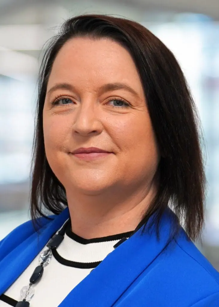 Woman with straight dark hair in a blue blazer and white top, smiling gently.