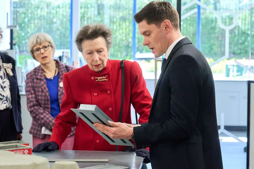 Man in suit shows tablet to Her Royal Highness, one in red coat, in modern office.