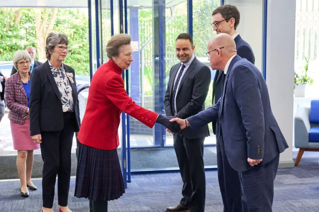 The Royal Highness The Princess Royal in red jacket shakes hands with suited man as others watch in an office.
