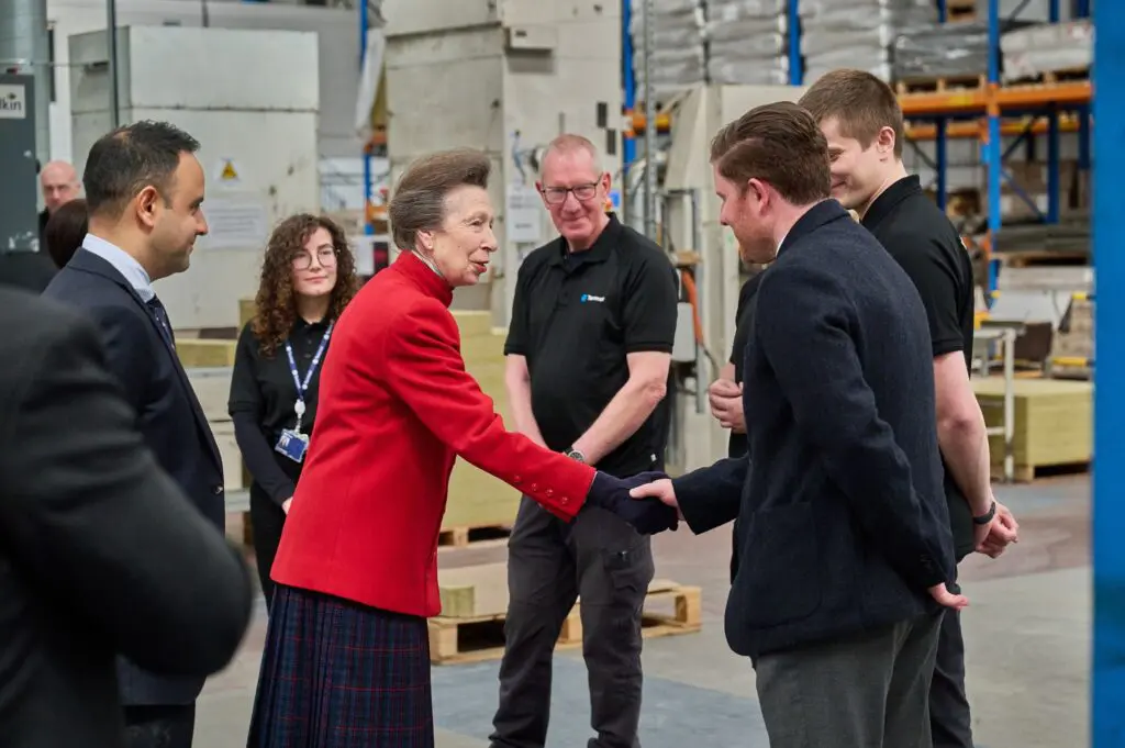 Woman in red jacket shakes hands with man in warehouse as others watch and smile.