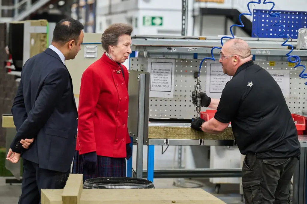 Three people in a workshop talk near machinery; one wears ear protection while working.