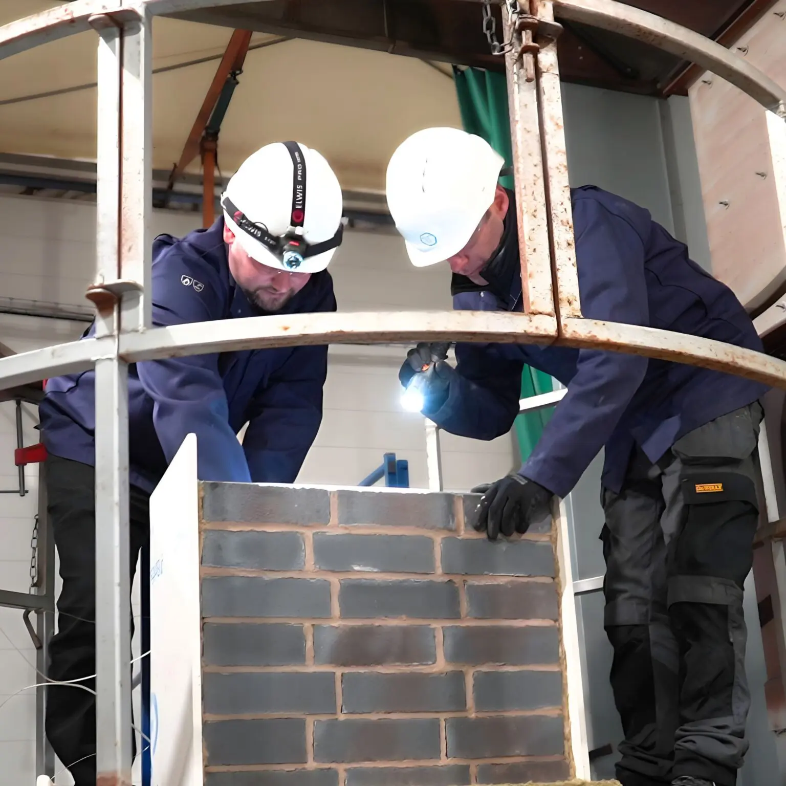 Two helmeted workers use a torch to inspect an indoor brick structure.