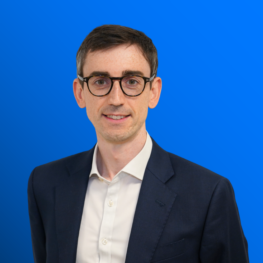 Smiling man in glasses and navy suit stands before a blue gradient background.
