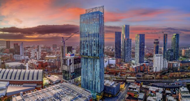 Manchester City centre Aerial night view of Deansgate Square and A city skyline at sunset with tall modern skyscrapers and cranes under a colorful sky.