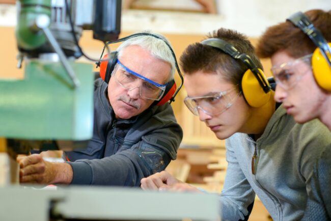 Woodwork,Apprenticeship An instructor teaches two students woodworking, all wearing safety glasses and ear protection.