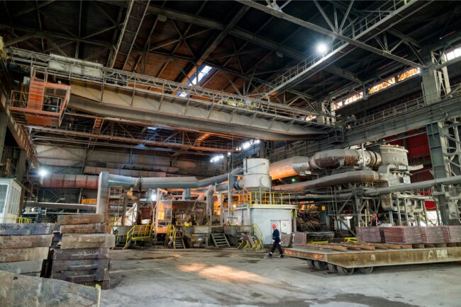 Large industrial factory interior with machinery, metal beams, and a worker walking near metal sheets.