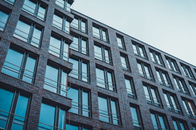 City,Housing,Constructed,Of,Dark,Brick,Exterior,And,Built,In Modern brick building with large glass windows and balconies, viewed from a low angle against a blue sky.