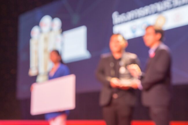 Disfocus,Of,The,Award,Ceremony,Theme,Creative.,Background,For,Business Three people on stage at an awards event, holding a trophy and a blank sign; background is blurred.