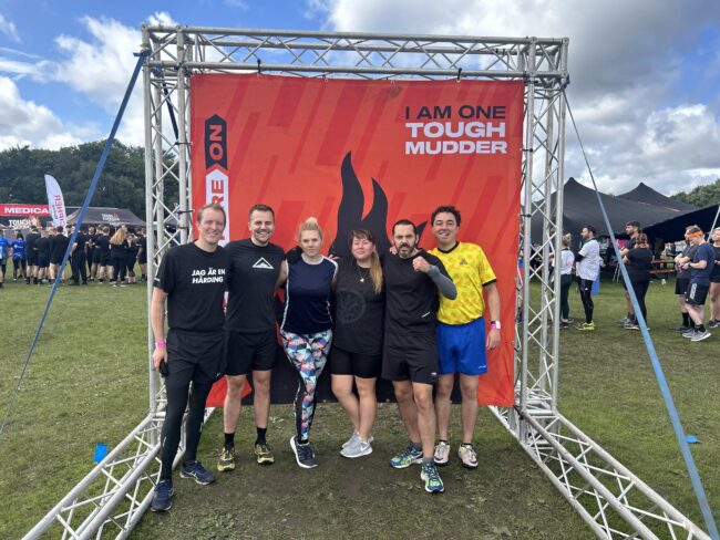 Six people in athletic wear pose and smile under a Tough Mudder event sign outdoors on a grassy field.