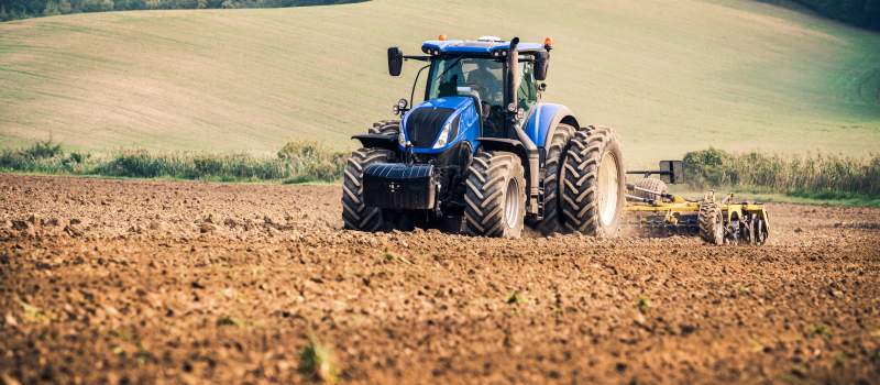 Blue tractor plowing a brown field with rolling green hills in the background under a clear sky.