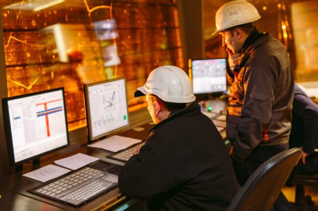 Two workers in hard hats monitor data on computers in an industrial control room with orange lighting.