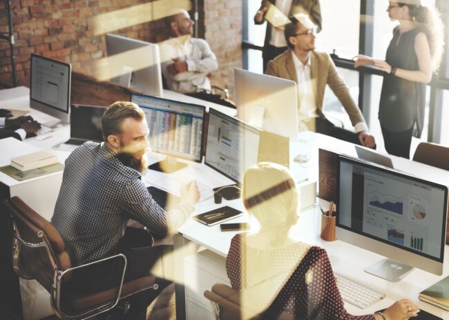 Office workers at desks using computers with charts; two people stand and talk in the background.