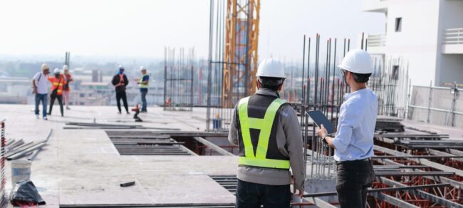 Construction workers in safety gear stand on a building site, discussing plans and observing progress.