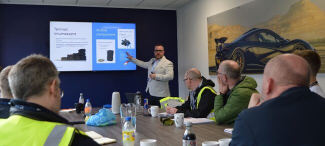 A man gives a presentation to a group seated at a table with a car photo and slideshow in the background.