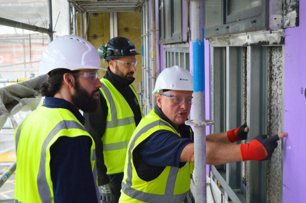 Three construction workers in safety gear talk by a purple wall at a building site.