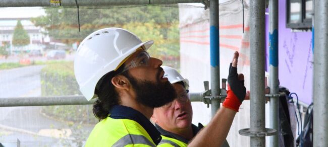 Two construction workers in hard hats inspect a building site; one points at a wall.