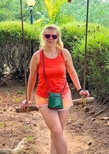 Woman in orange tank top and shorts sits on an outdoor swing, sunglasses on, green purse by her side.