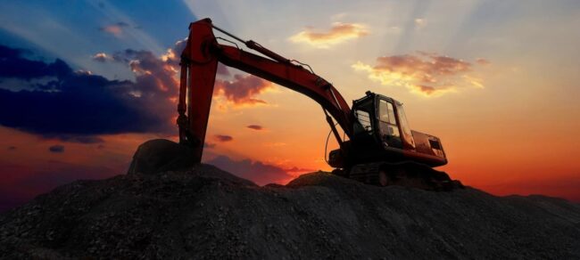 Excavator silhouetted on a dirt mound against a vibrant, cloudy sunset sky.