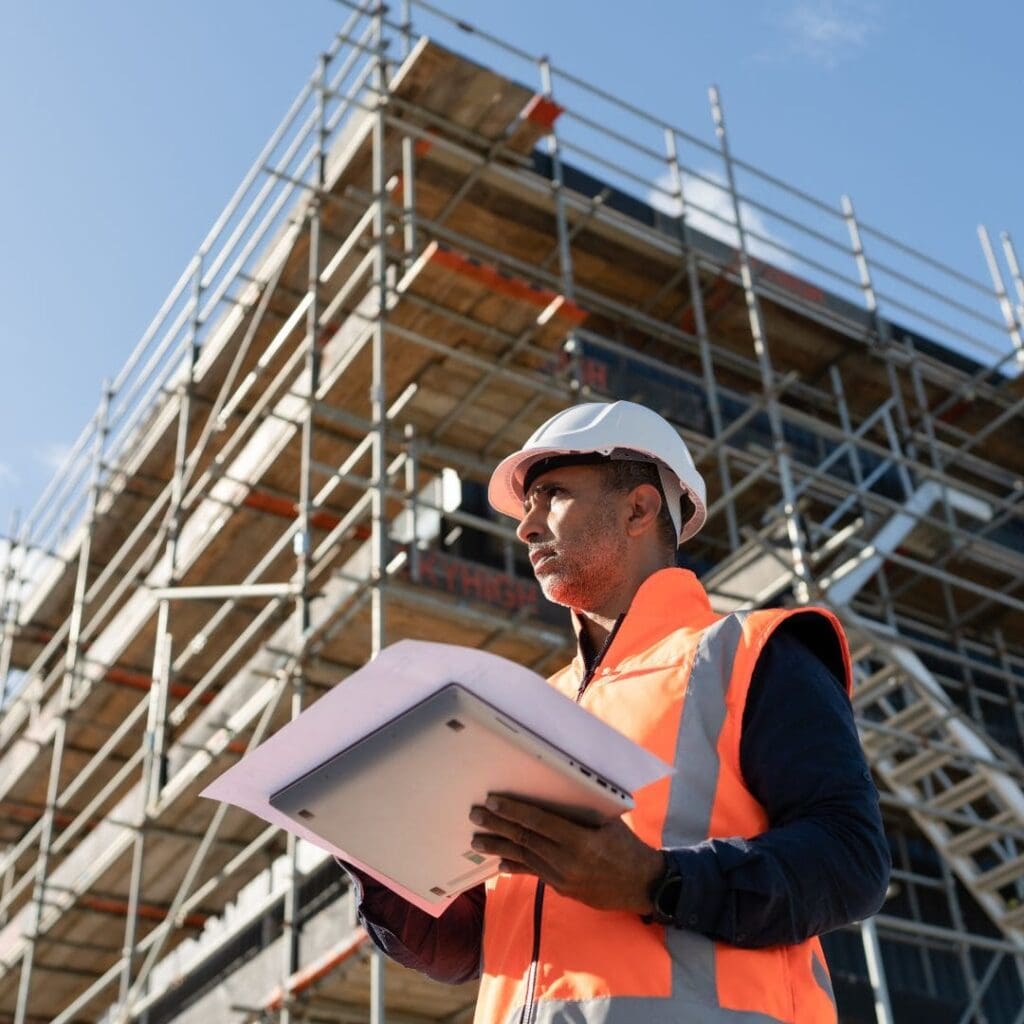 Man in safety vest and helmet holds documents before scaffolding on a clear day.