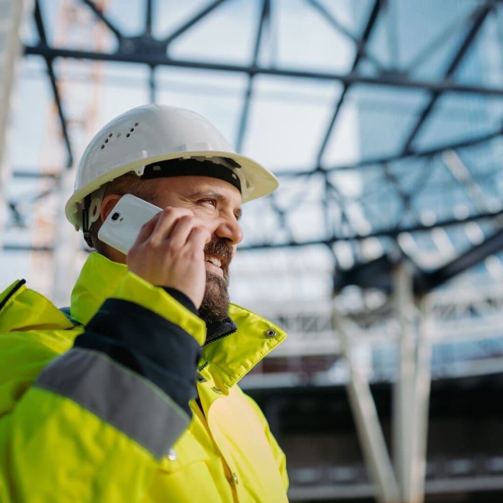 Construction worker in hard hat and yellow jacket talks on smartphone at building site.