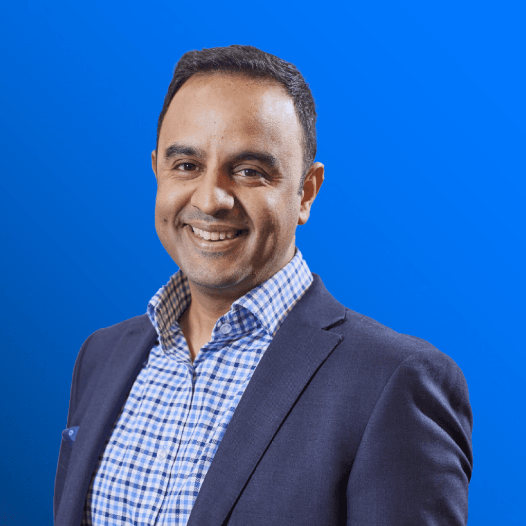 Man in a blazer and checkered shirt smiling in front of a bright blue background.