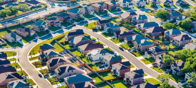 Aerial shot of a suburban area with houses, green lawns, and leafy streets.
