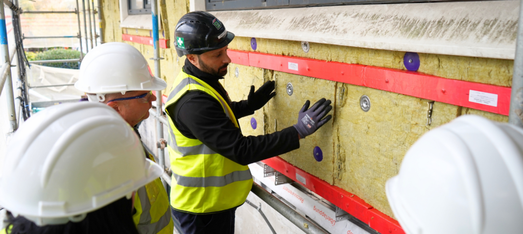 Workers in safety gear discuss and inspect wall insulation at a construction site.