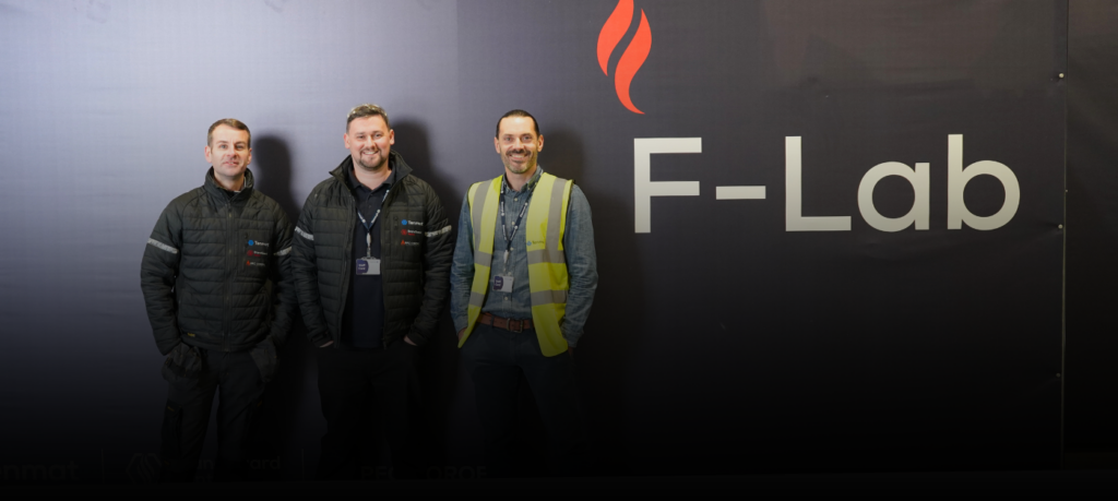 Three men stand before a black wall with a flame logo and “F-Lab.” written on it.