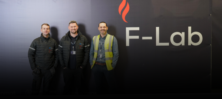 Three men stand before a black wall with a flame logo and “F-Lab.” written on it.