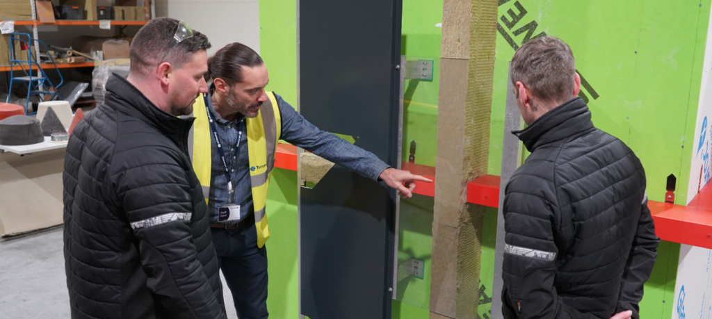 Three men discuss a glass wall in an industrial workplace.