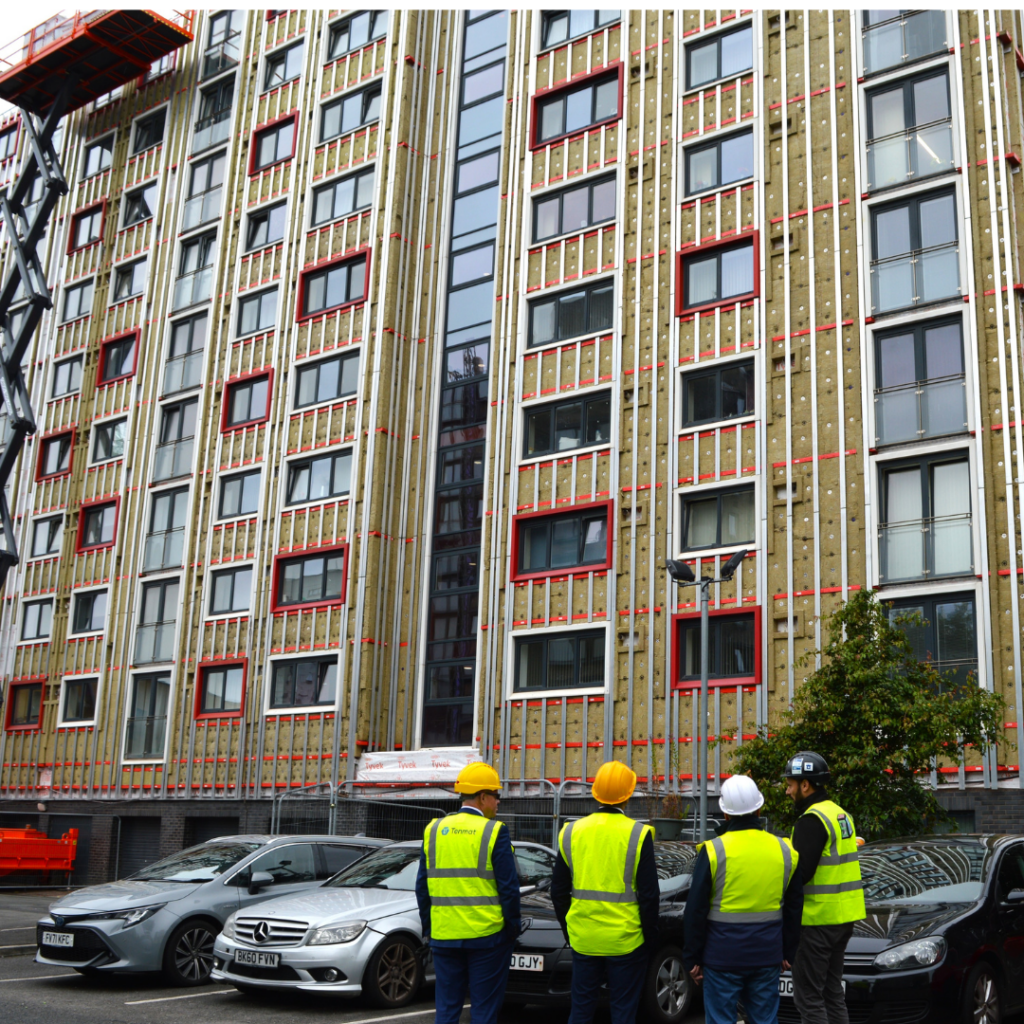 Four workers in safety gear watch a tall building being constructed or renovated.