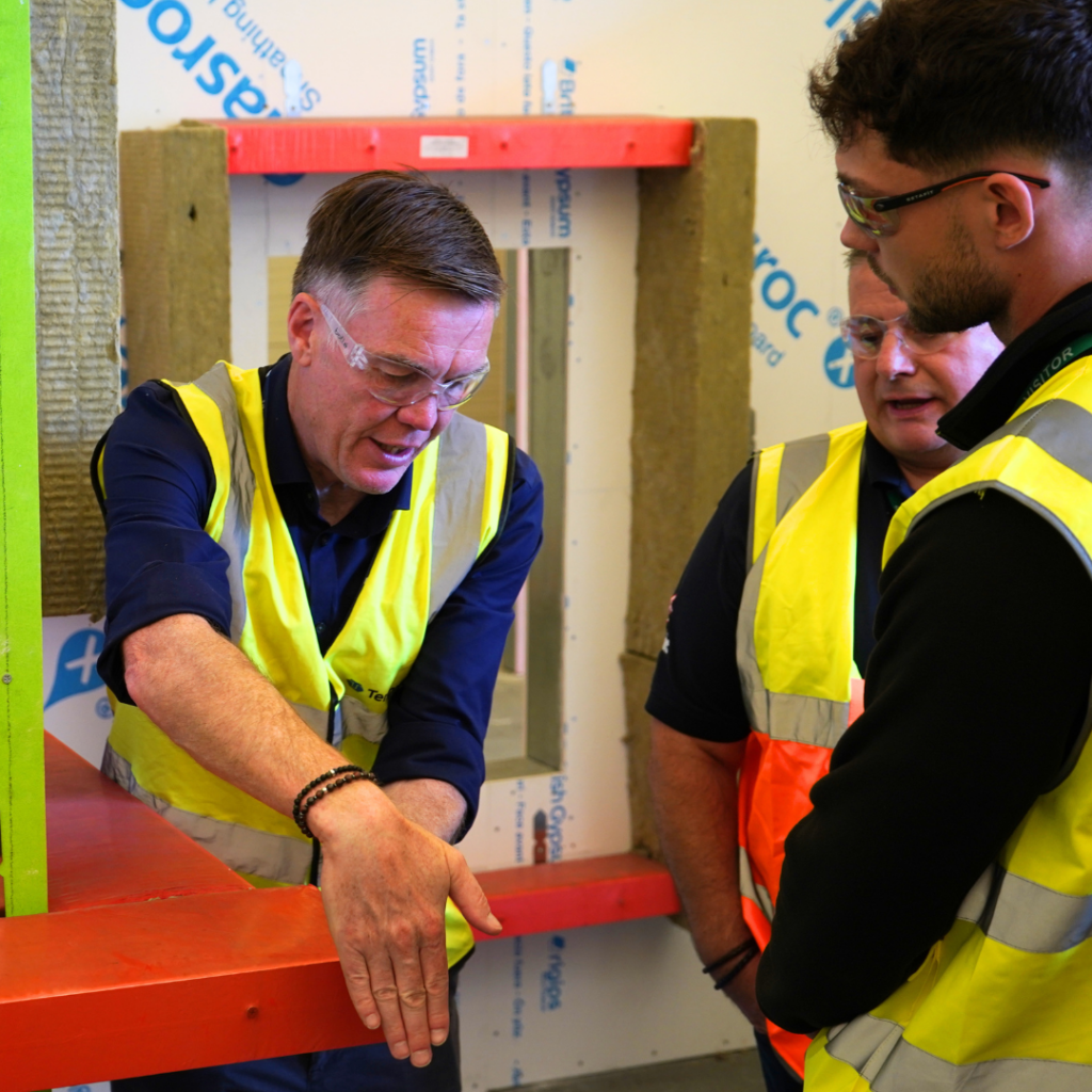 Three men in safety vests talk over materials at an indoor building site.