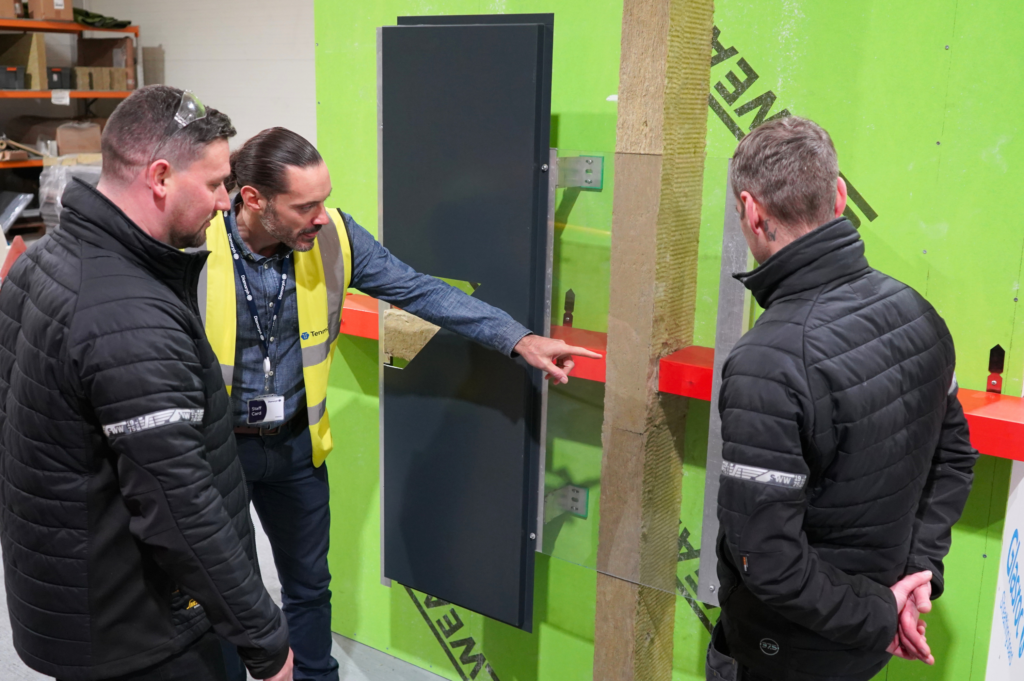 Three men inspect a panel and insulation on a green wall in an industrial area.