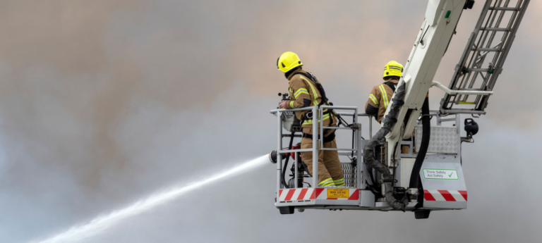 Two firefighters on a lift fight smoke as an evacuation alert system blares nearby.