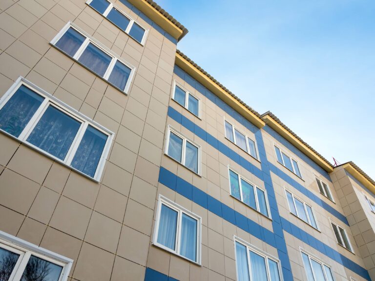 Tall beige and blue block of flats with many windows, seen from below against the sky.