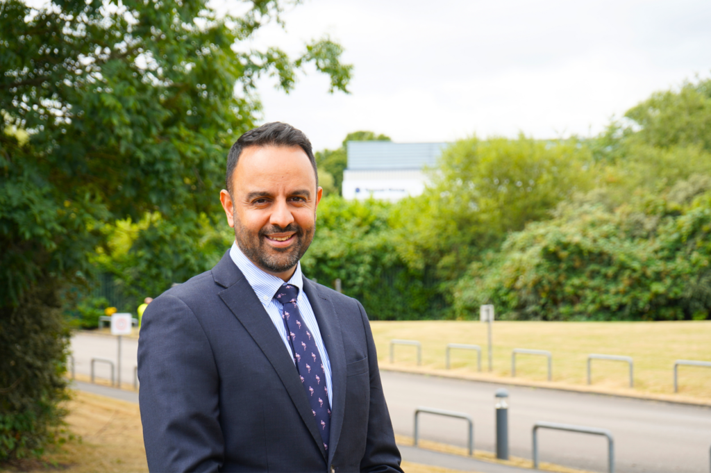 Smiling man in a suit outside, with trees and a road behind him.