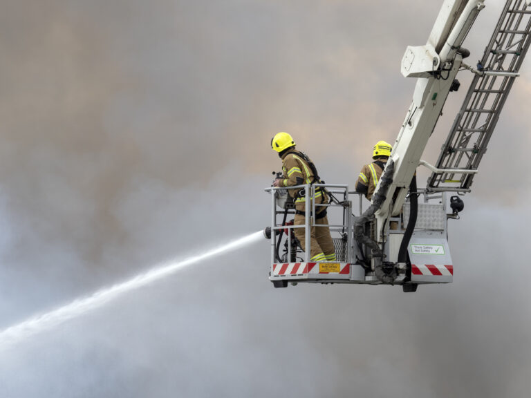 Firefighters on a ladder spray water into thick smoke.