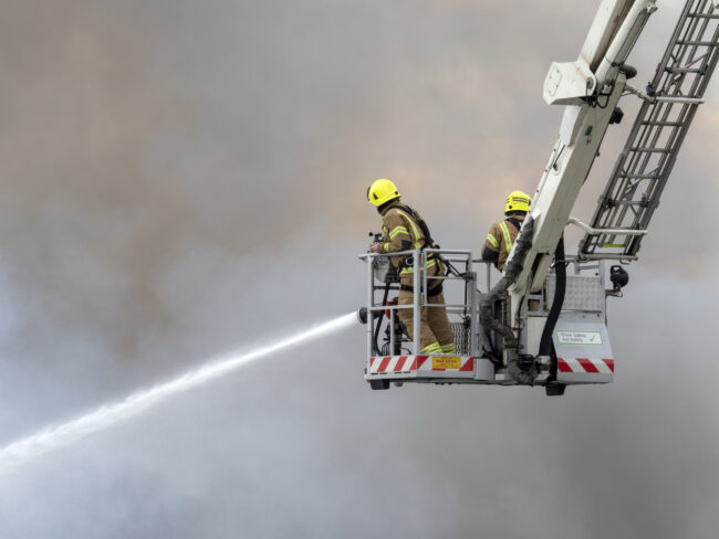 Firefighters on a ladder spray water into thick smoke.