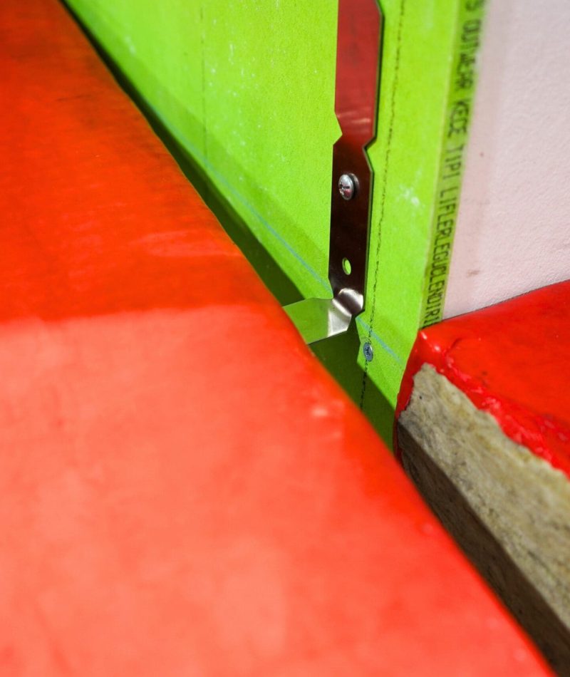 Metal corner bead on green drywall beside a red construction surface, close-up view.
