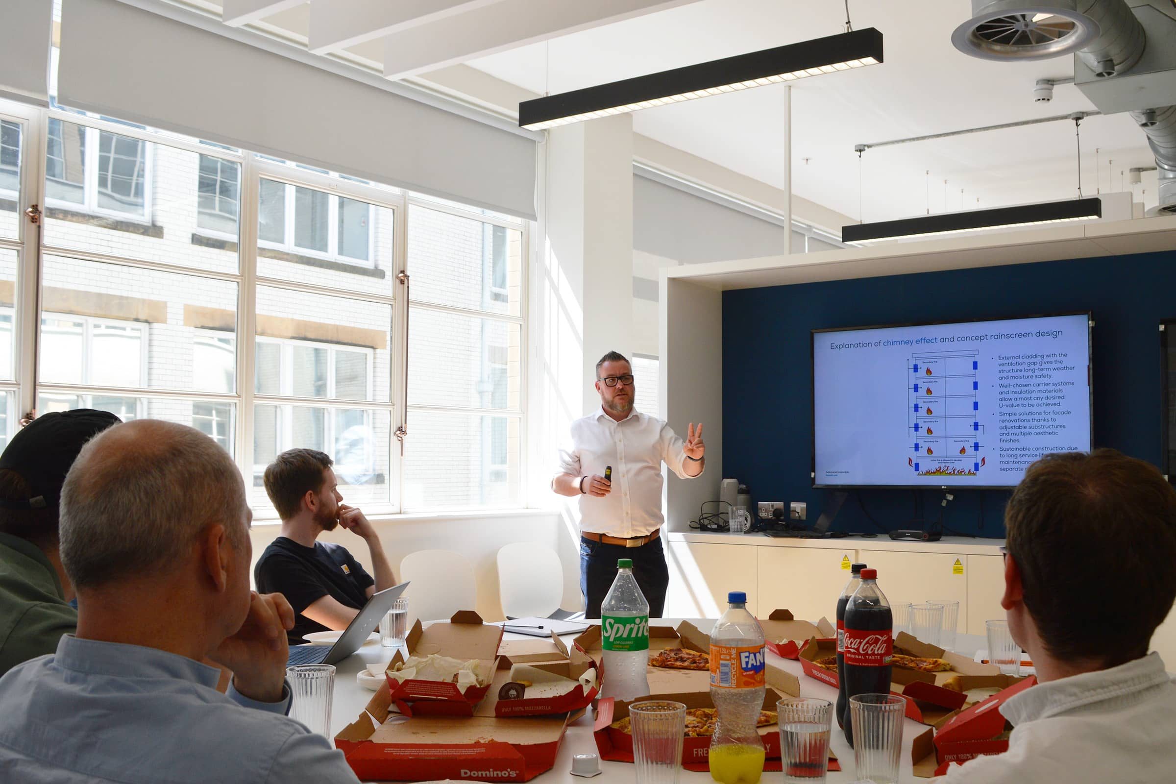 Man shows a fire protection chart to colleagues in a meeting room with pizza and drinks.