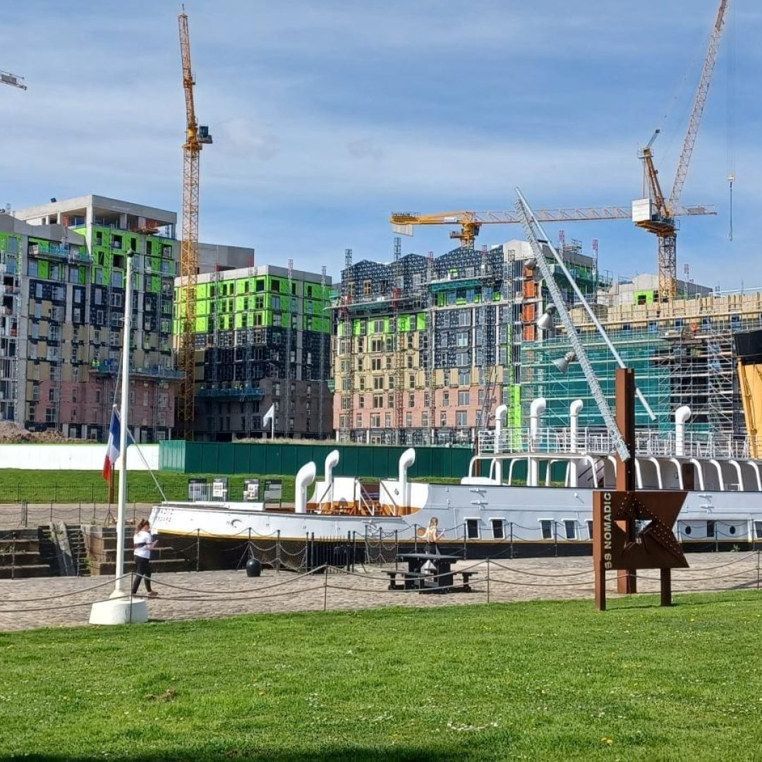 A historic ship docks by modern buildings and cranes under construction.